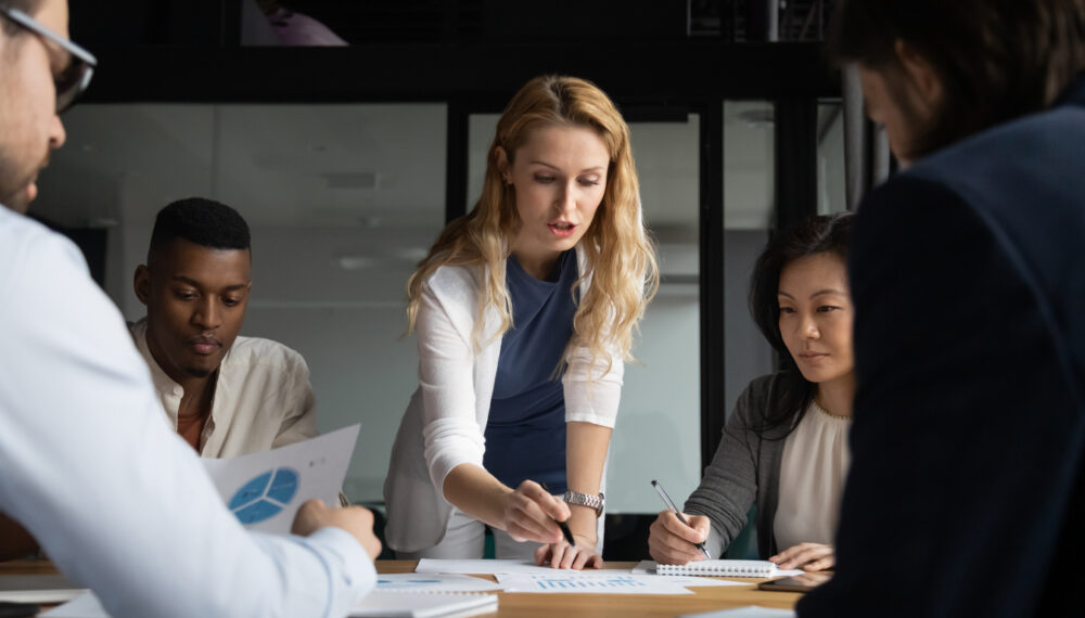 Concentrated young businesswoman explaining market research results in graphs to mixed race colleagues. Focused group of diverse employees holding brainstorming meeting, discussing project ideas.